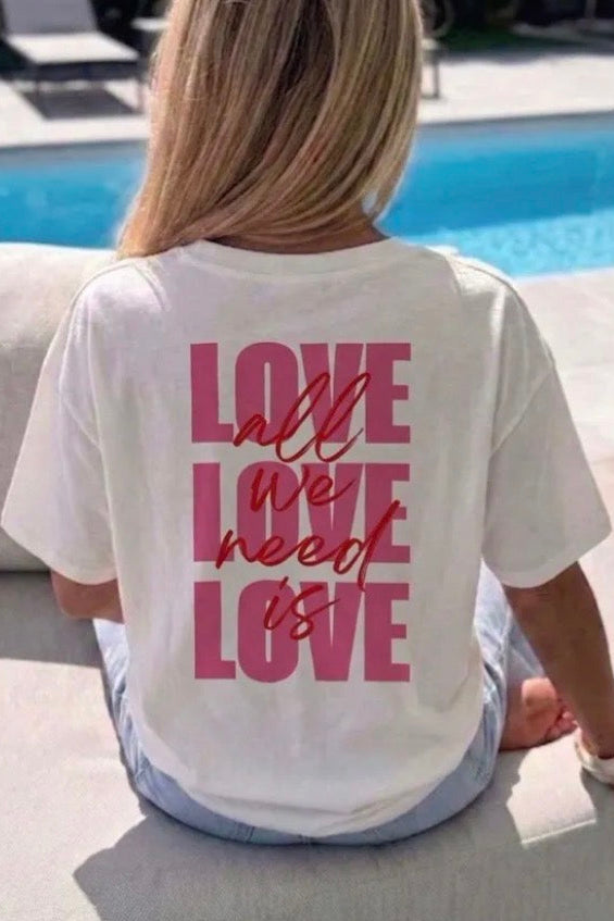 A woman sitting outdoors by a swimming pool, viewed from behind, wearing a white t-shirt with a pink graphic text design on the back that reads 'LOVE all we need is LOVE'.