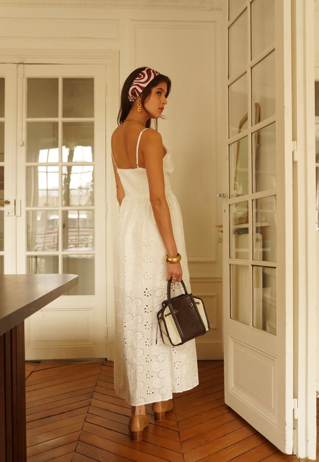 A woman stands in a bright room with white paneled doors and wood floors, wearing a white eyelet midi dress with spaghetti straps and holding a black handbag.