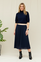 A woman wearing a green funnel neck jumper with short sleeves, paired with blue jeans and brown ankle boots. She has sunglasses on and is standing against a white wall with a yellow leaf on the ground.