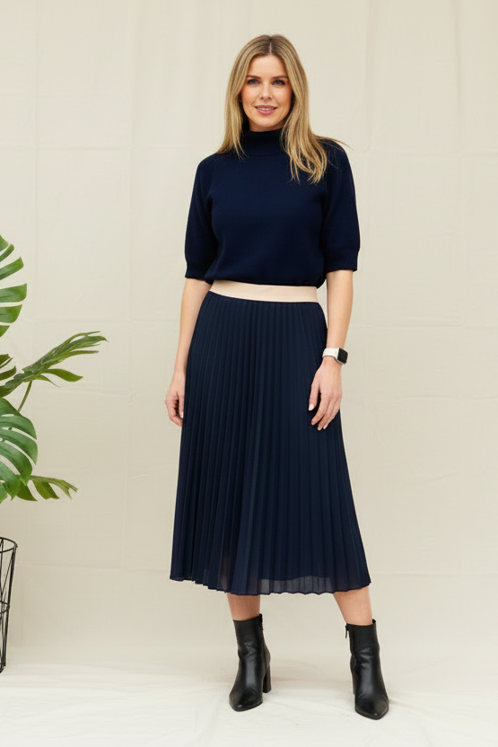 A woman wearing a green funnel neck jumper with short sleeves, paired with blue jeans and brown ankle boots. She has sunglasses on and is standing against a white wall with a yellow leaf on the ground.