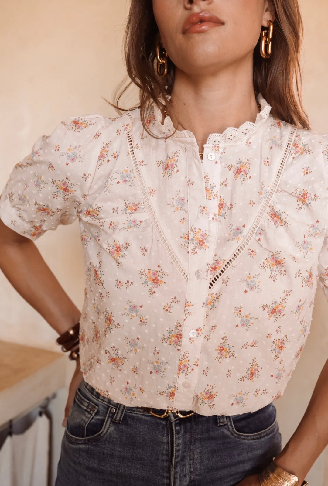A woman wearing a short-sleeve white eyelet blouse with a delicate multicolor floral print and lace-up front, paired with dark blue jeans.