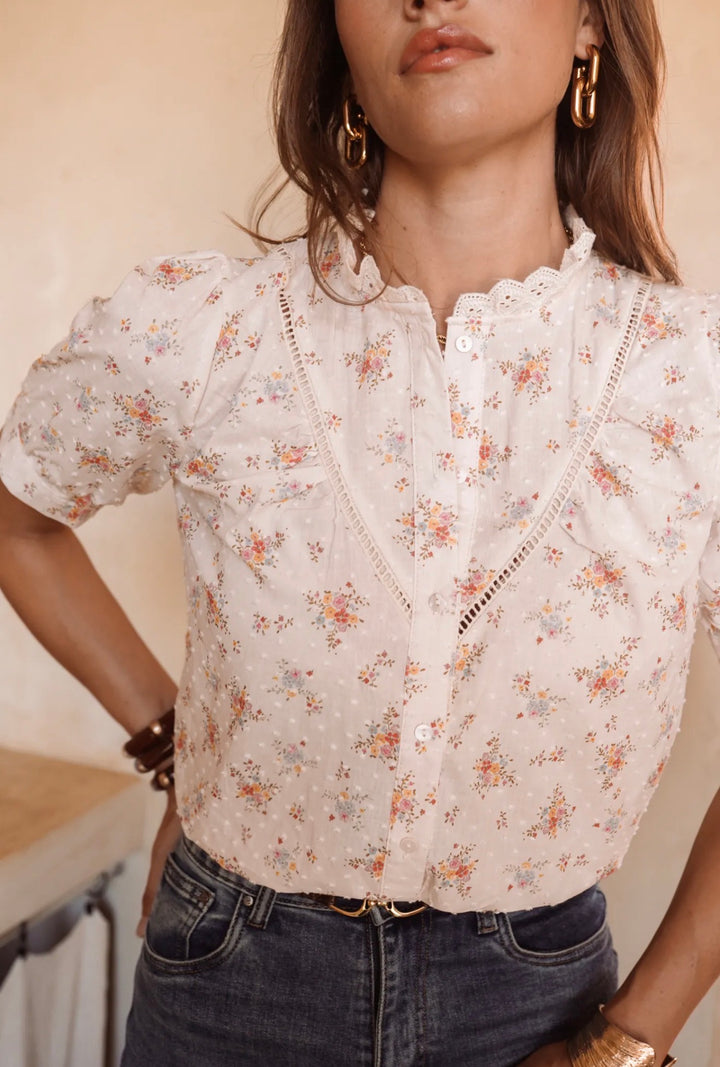 A woman wearing a short-sleeve white eyelet blouse with a delicate multicolor floral print and lace-up front, paired with dark blue jeans.