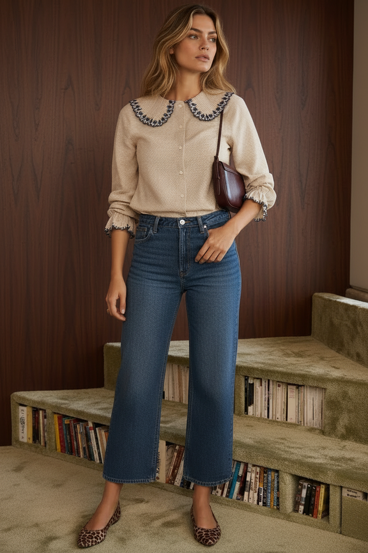 A woman sits on a textured green bench wearing a beige gingham button-up blouse with a large navy and white embroidered Peter Pan collar, paired with blue jeans and a dark brown shoulder bag.