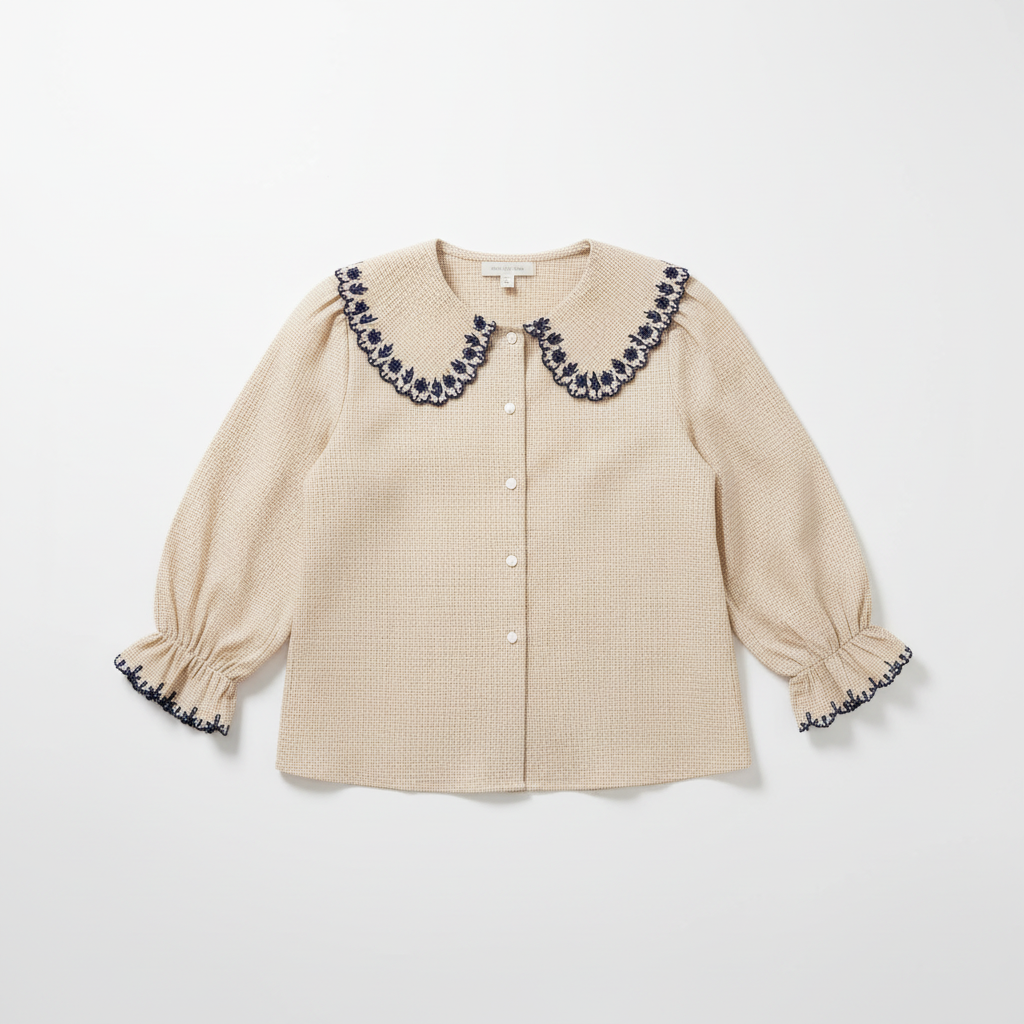 A woman sits on a textured green bench wearing a beige gingham button-up blouse with a large navy and white embroidered Peter Pan collar, paired with blue jeans and a dark brown shoulder bag.