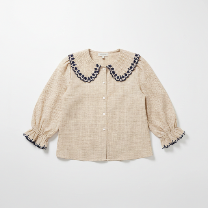 A woman sits on a textured green bench wearing a beige gingham button-up blouse with a large navy and white embroidered Peter Pan collar, paired with blue jeans and a dark brown shoulder bag.