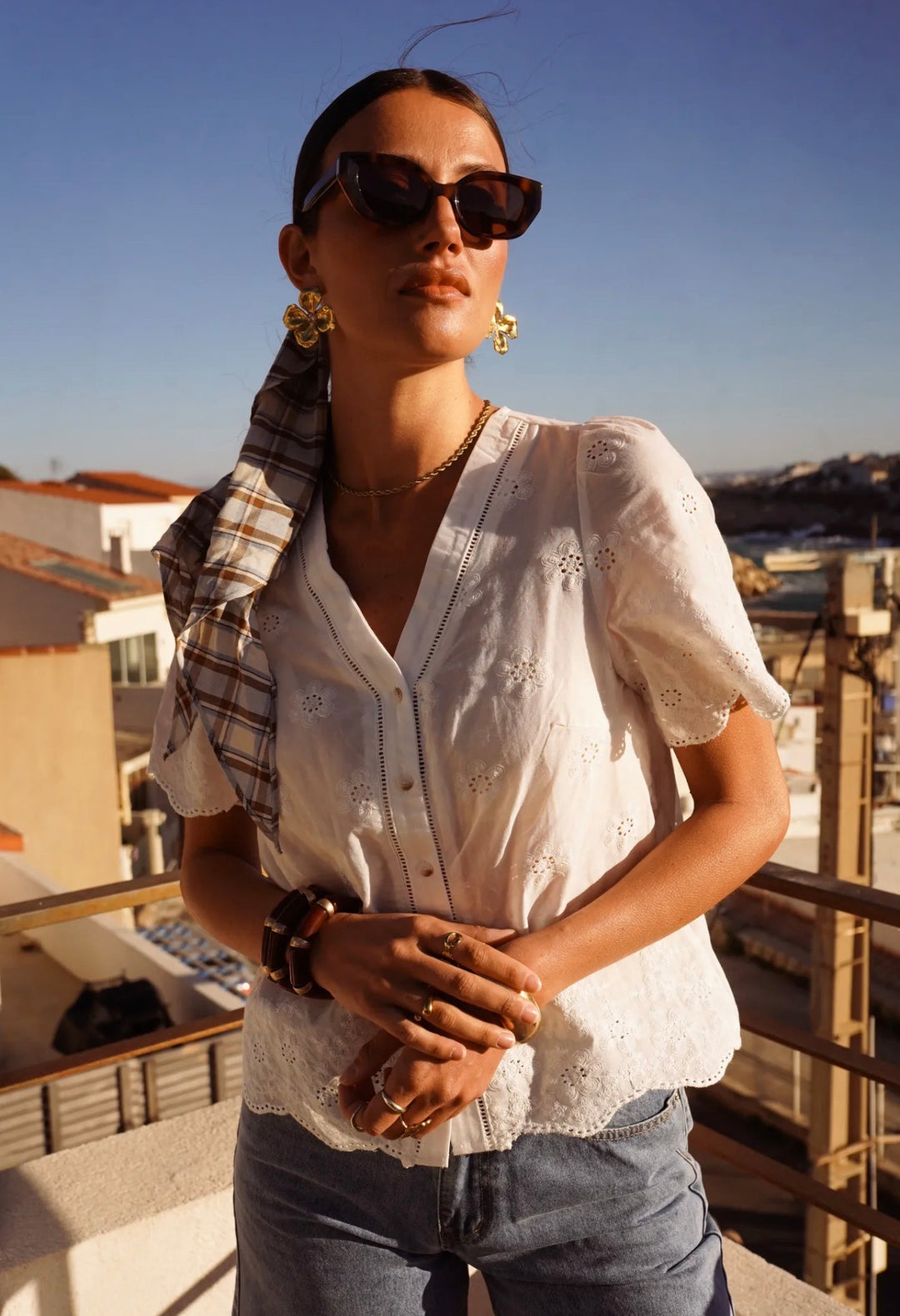A woman wearing a white embroidered short-sleeve blouse, blue jeans, sunglasses, and a plaid scarf stands on a balcony with a city and sea background under a clear blue sky.