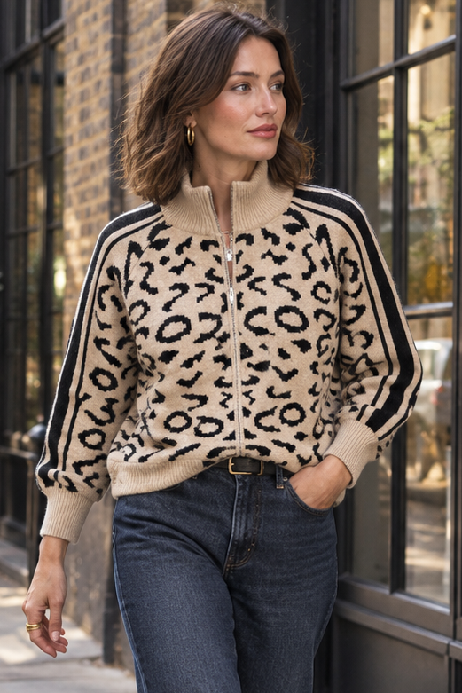 A woman walking on a city sidewalk wearing a beige and black leopard-print zip-up sweater with black stripes on the sleeves, dark blue jeans, and white sneakers.