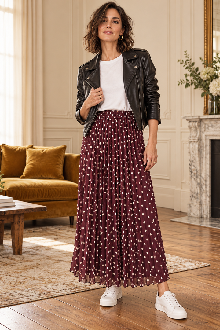 A woman stands in a bright living room wearing a black leather jacket, a white crew-neck top, and a long burgundy pleated skirt with small white polka dots. She is also wearing white sneakers.