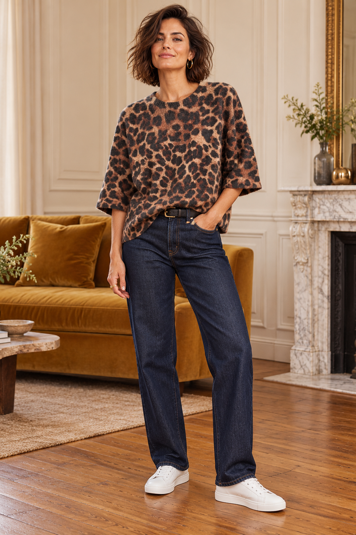 A woman stands in a living room wearing a brown and black leopard-print blouse, dark blue straight-leg jeans, and white sneakers.
