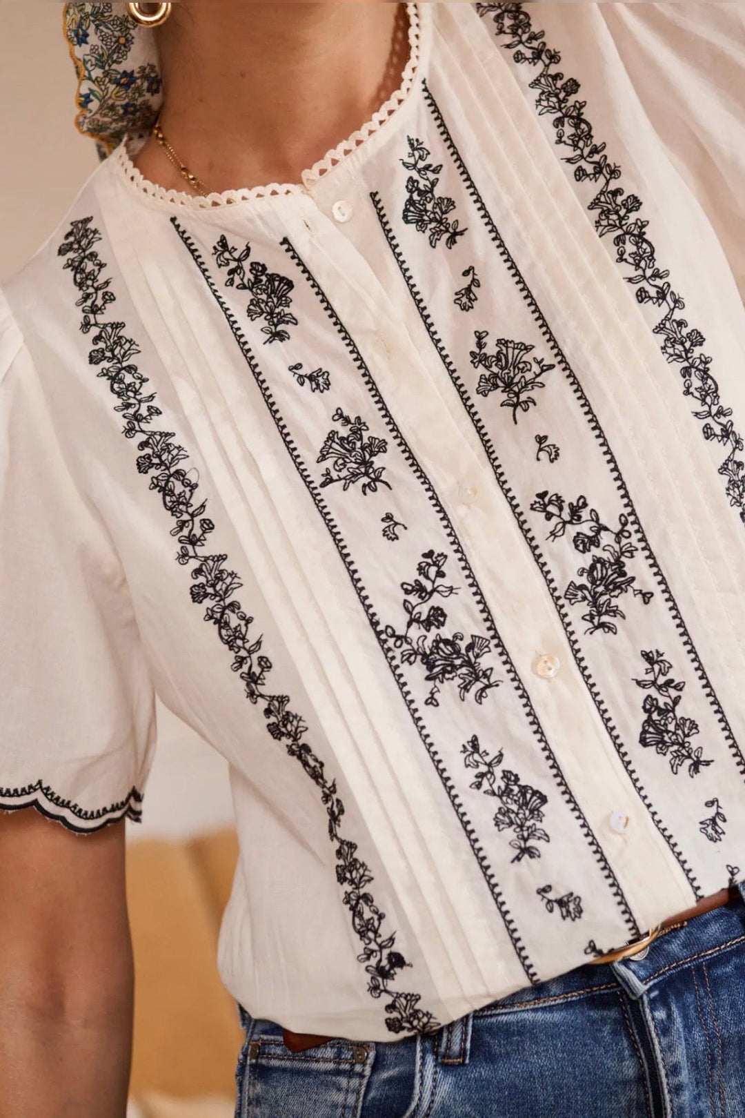 White blouse with black floral embroidery worn by a person, blurred background