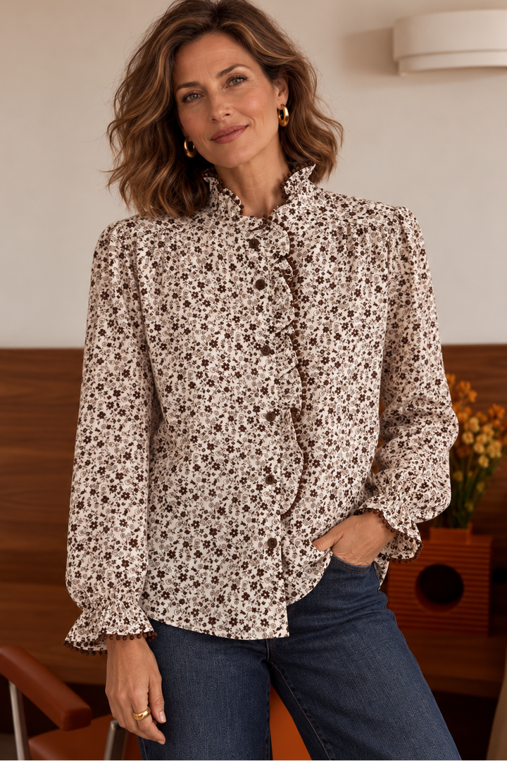 A woman wearing a beige long-sleeve blouse with a dense black floral print, paired with dark blue jeans, standing in a warm-toned indoor setting.