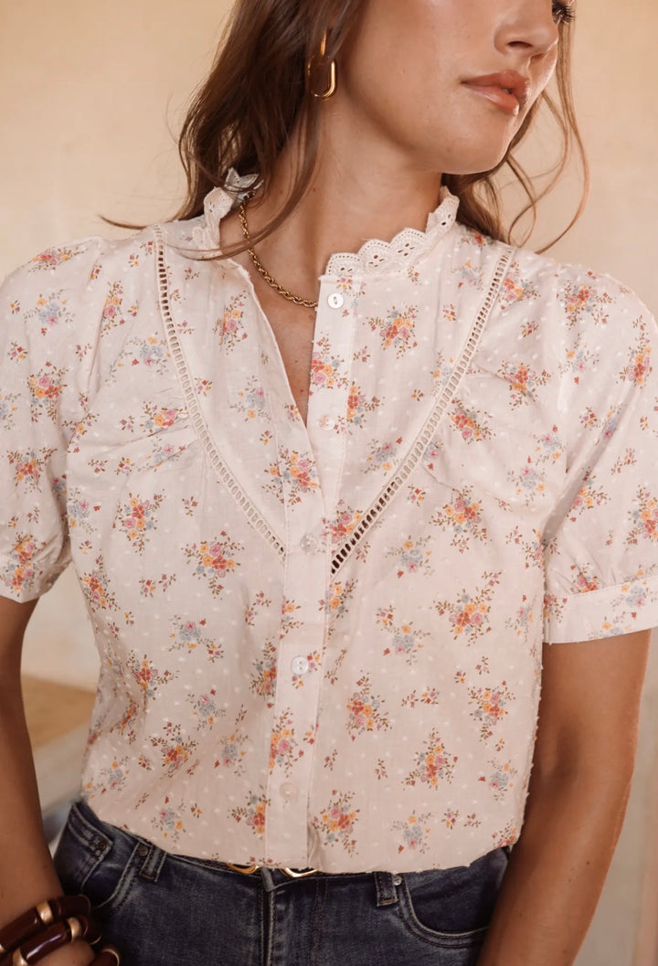 A woman wearing a short-sleeve button-front blouse with a delicate floral print, lace trim, and scalloped collar, paired with dark blue jeans against a warm beige background.