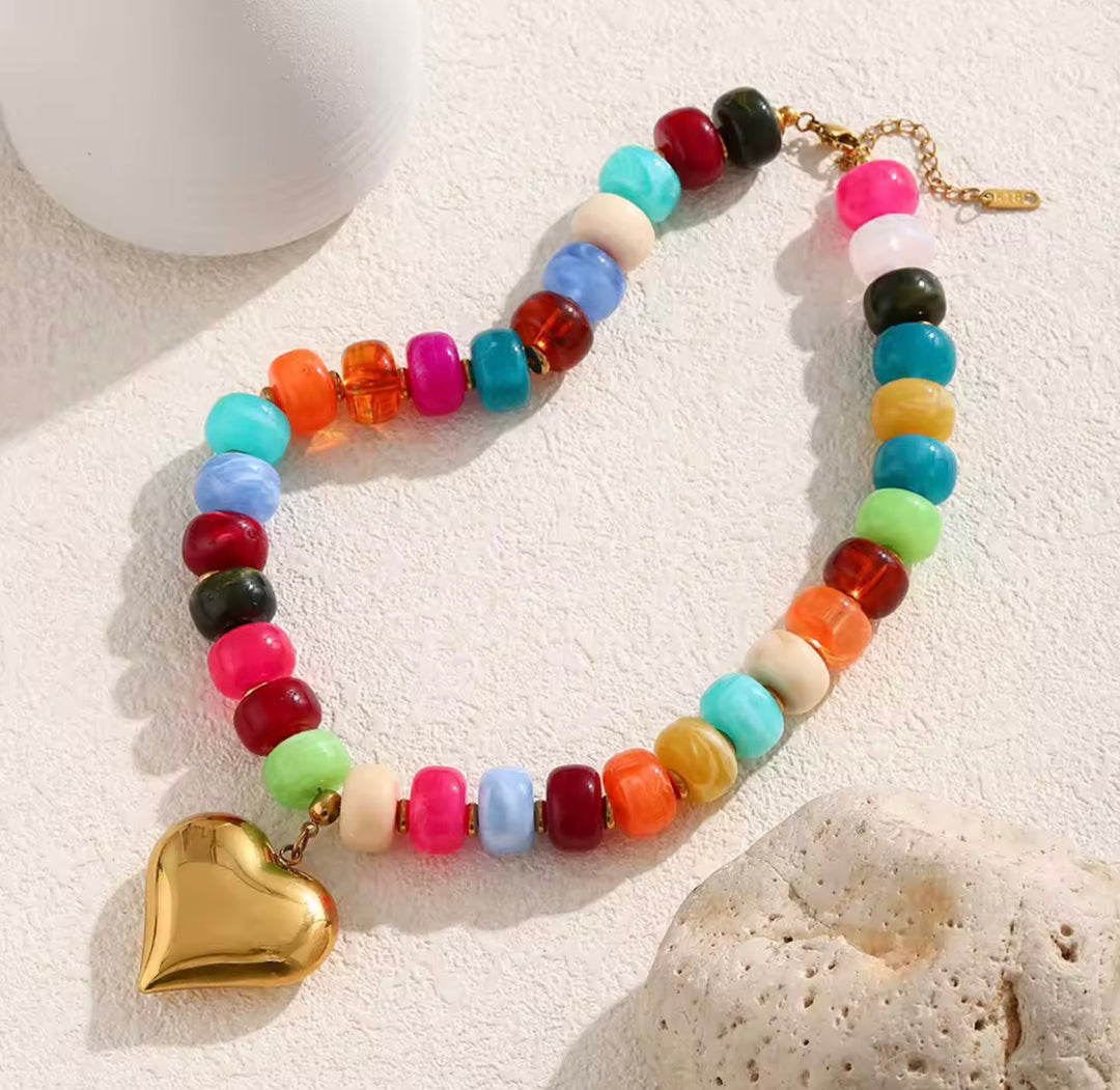 A colorful beaded necklace with a gold heart pendant, displayed on a textured white surface with decorative stones.