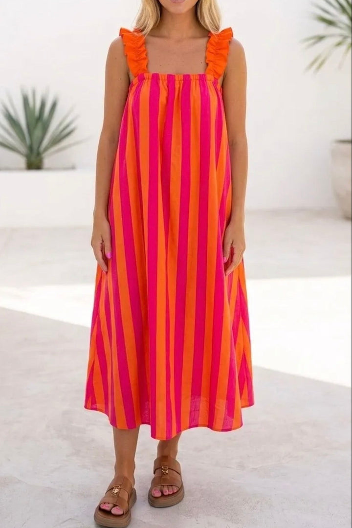 A woman modeling a sleeveless midi dress with vertical orange and pink stripes and ruffled shoulder straps, standing in a bright, minimalist white indoor-outdoor space with potted plants.