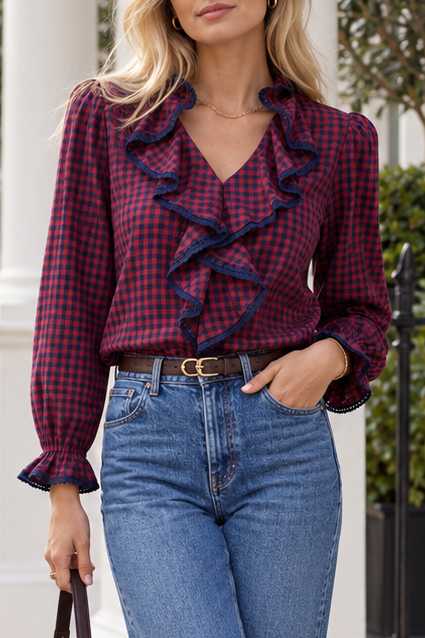A woman wearing a long-sleeve red and navy blue gingham blouse with dramatic ruffles down the front and ruffled cuffs, paired with blue jeans and a brown belt.