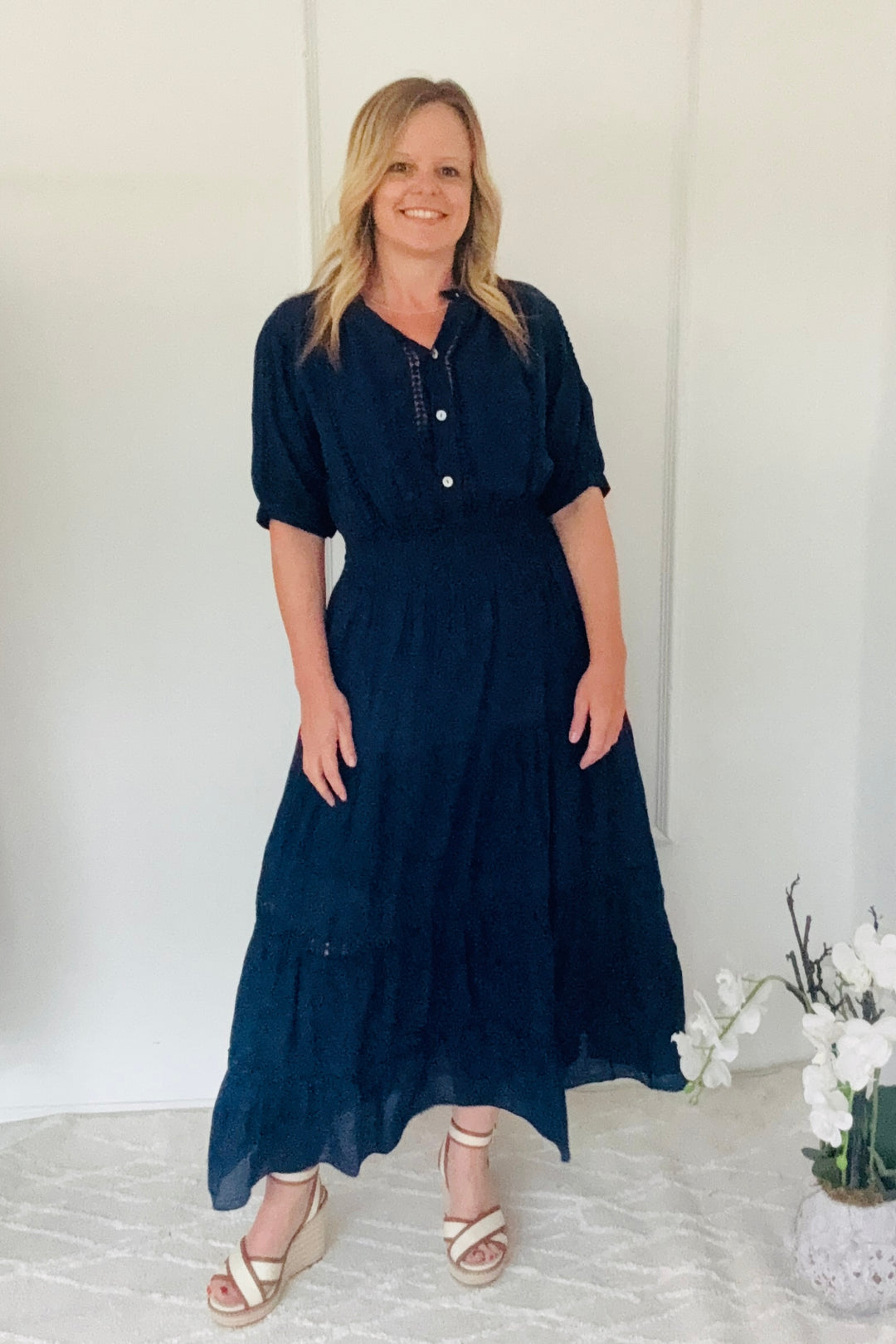 Woman wearing a navy dress standing against a white background with a vase of flowers.