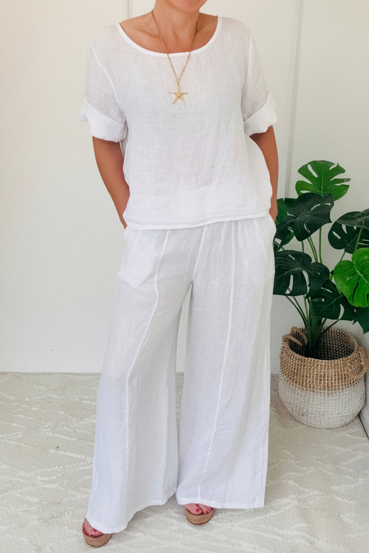 Person wearing a white outfit standing in a minimal indoor setting with a plant in the background.