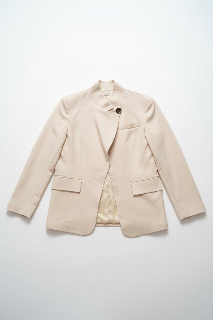 A light beige wrap-style blazer with a single dark button closure, displayed on a hanger on a clothing rack inside a retail store.