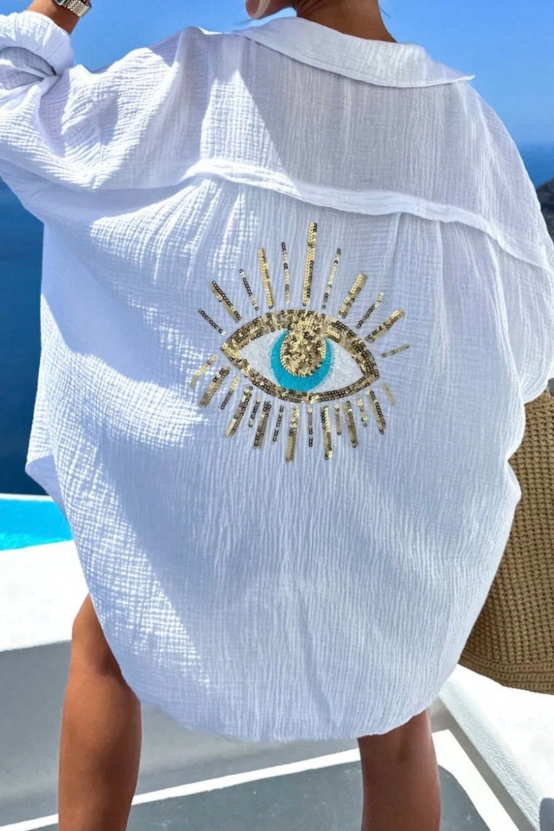 Woman in a white cover-up with an embroidered design, standing on a scenic location with blue sky and ocean.