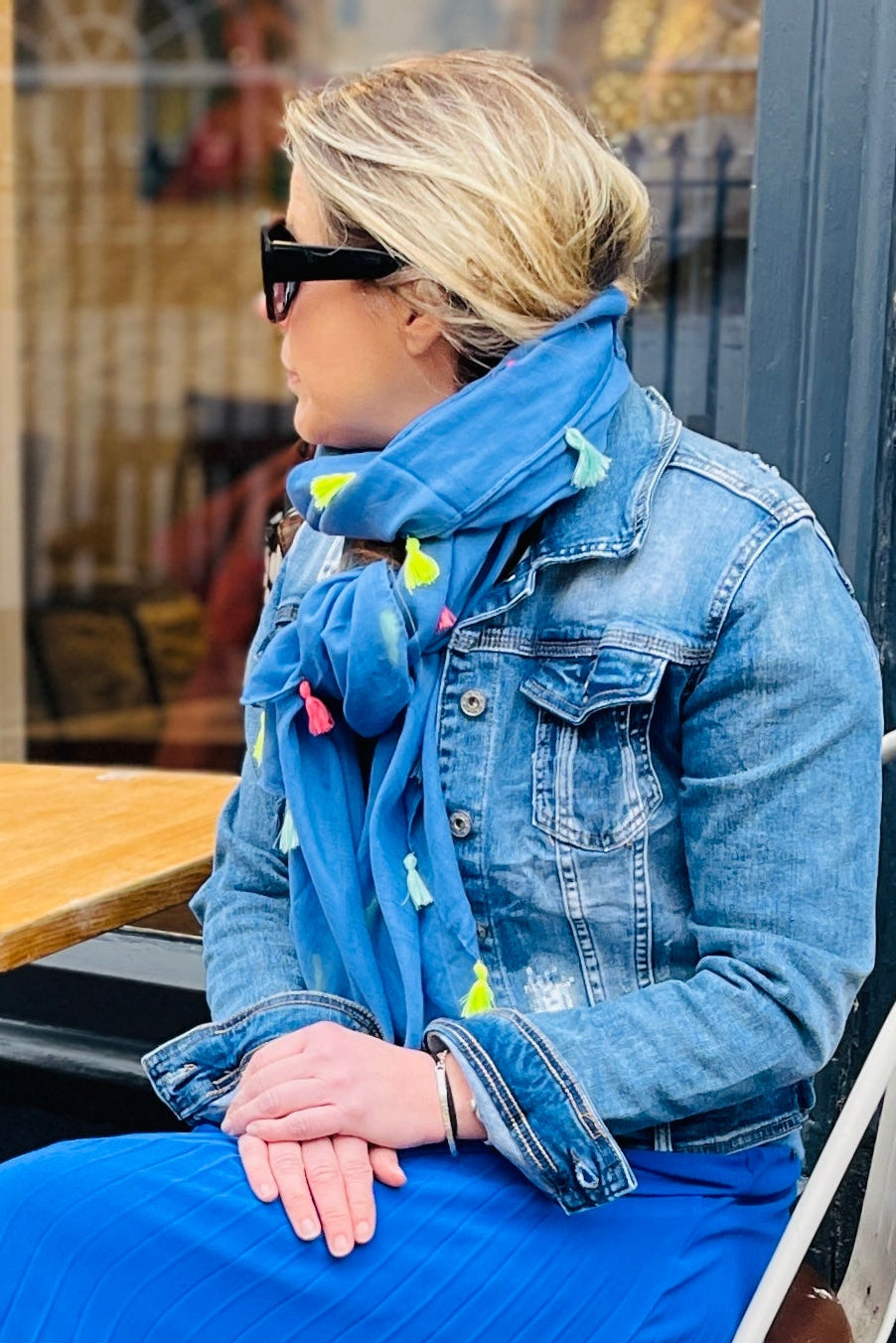 A person seated at an outdoor cafe wearing a royal blue pleated maxi skirt with an elasticated waistband, paired with a denim jacket and white shoes.