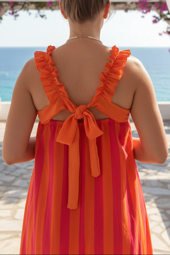 A woman viewed from behind wearing a sleeveless orange dress with ruffled straps and a bow at the back, standing in a clothing store.