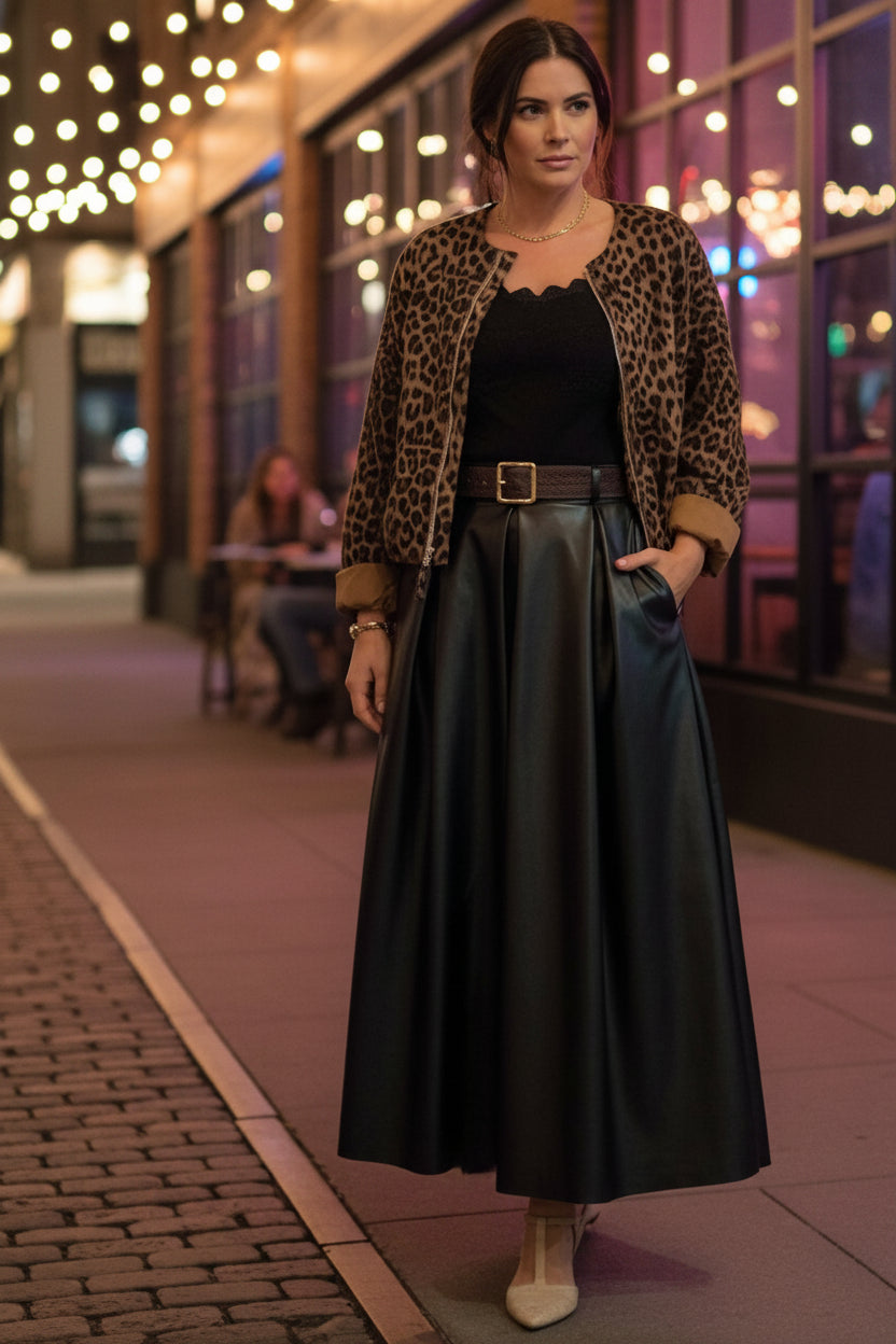 A person wearing a leopard-print zip-up jacket with tan cuffs, a black velvet top, a long black skirt, and beige flats, standing in front of a paneled wall.