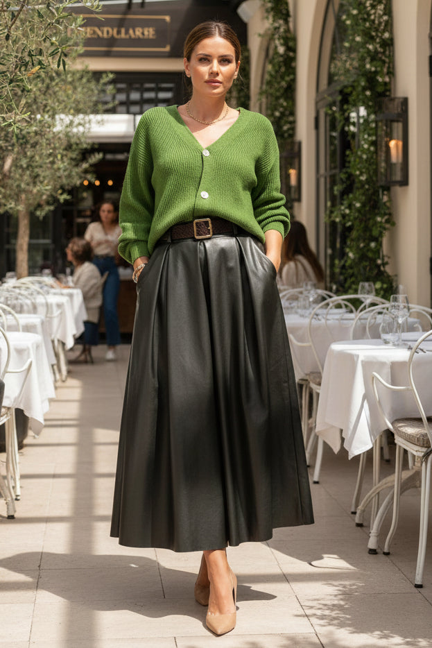A person wearing a green knit cardigan and a long black pleated skirt with a black belt, standing indoors against a paneled wall.