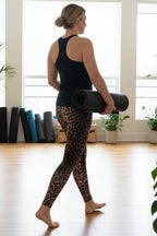 Woman in leopard print leggings and black tank top holding a yoga mat in a room with large windows and plants.