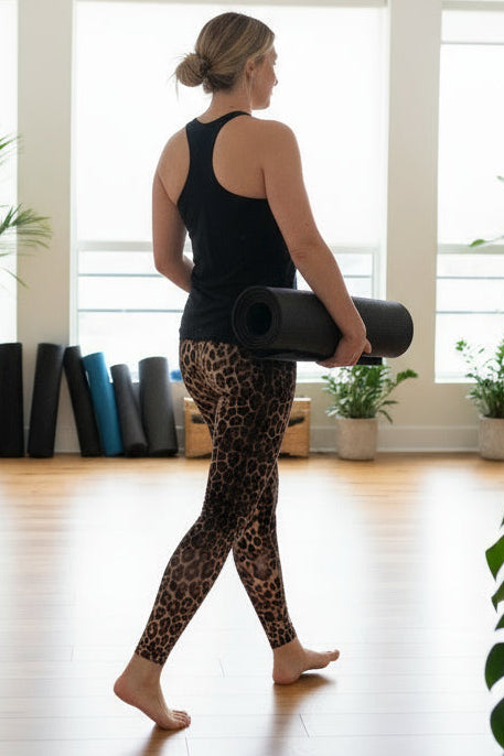Woman in leopard print leggings and black tank top holding a yoga mat in a room with large windows and plants.