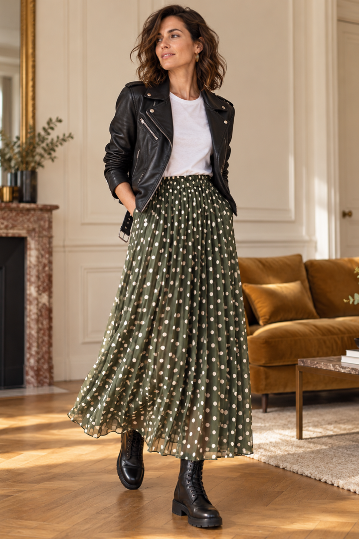 A woman stands in a bright, elegant room wearing a black leather moto jacket, a white crewneck t-shirt, and a high-waisted olive green pleated midi skirt with white polka dots. She is also wearing black leather ankle boots.