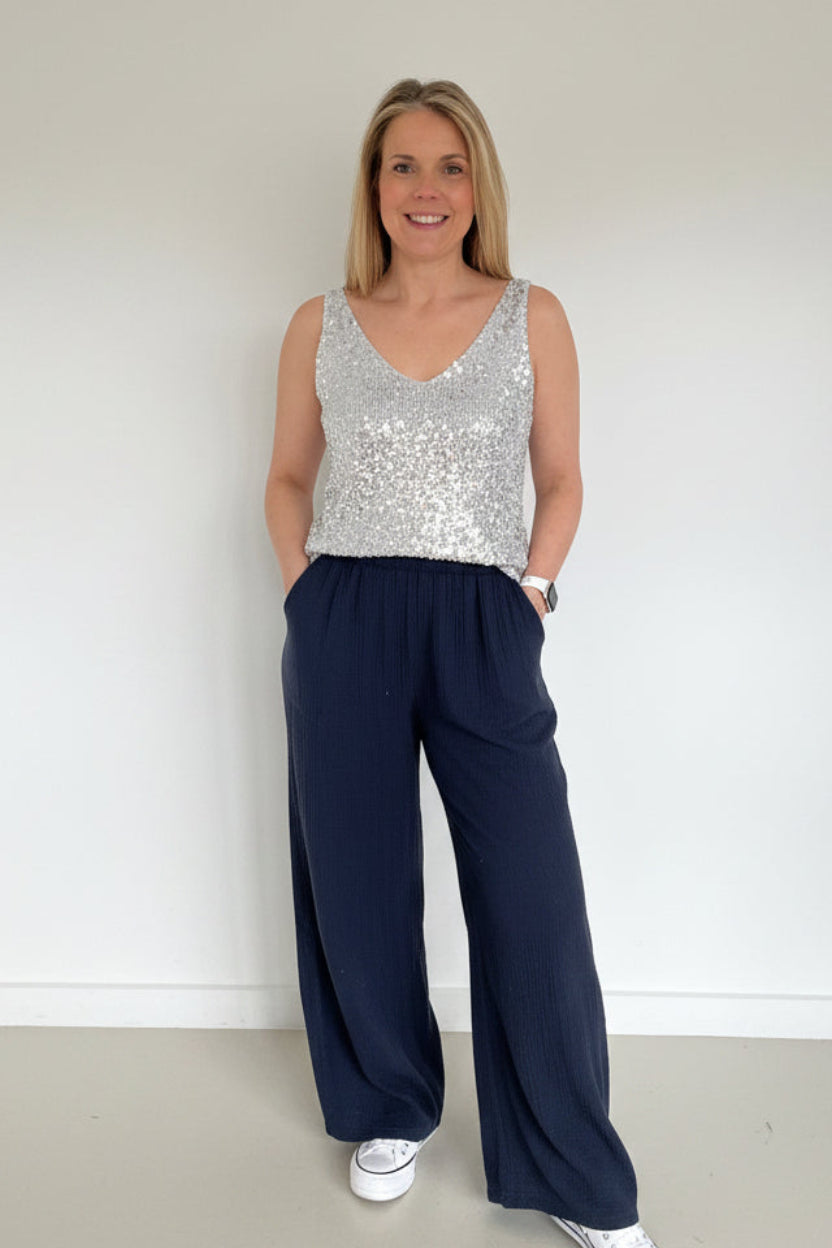 Person wearing a sleeveless top and navy pants standing in a room with a white wall and a rug.
