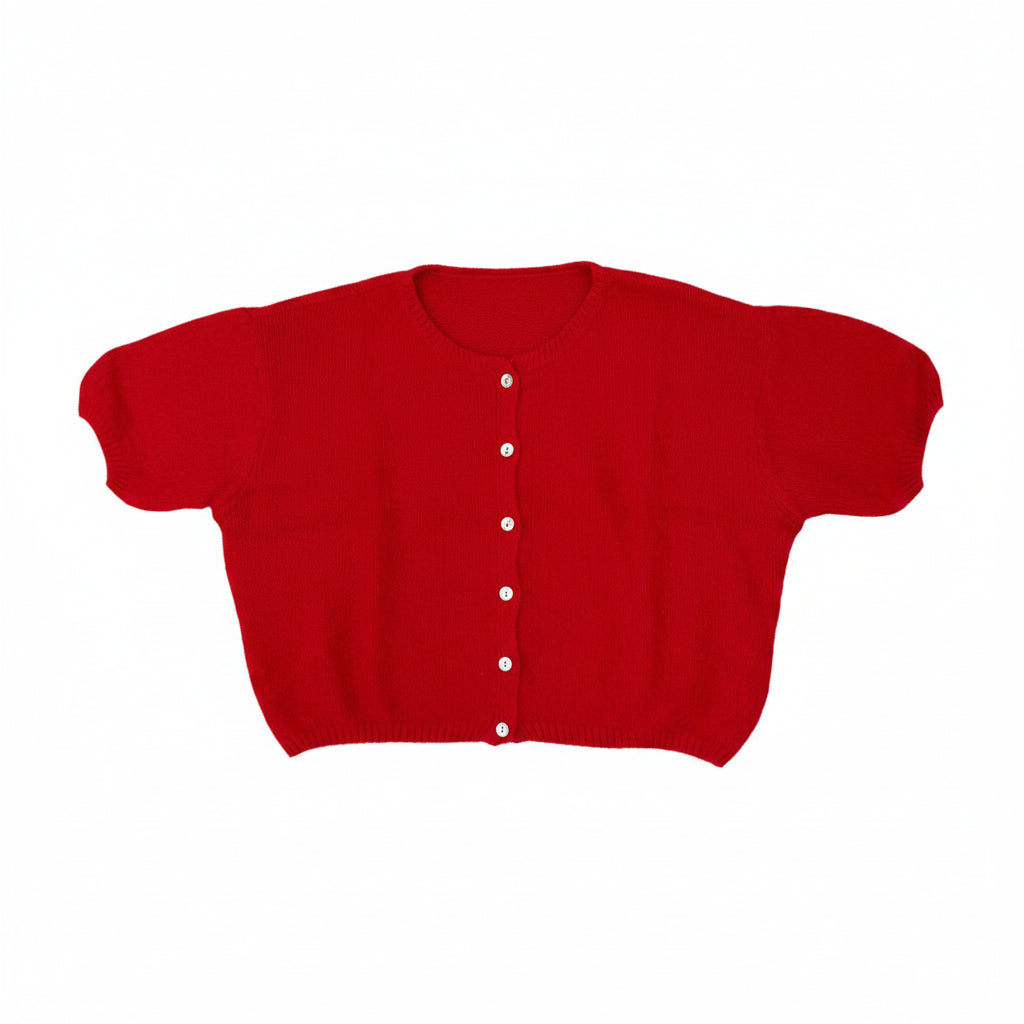 A bright red short-sleeve cropped cardigan sweater with white buttons, laid flat on a gray carpet. A person's feet in beige shoes are visible at the bottom edge.