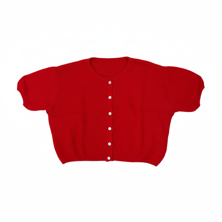 A bright red short-sleeve cropped cardigan sweater with white buttons, laid flat on a gray carpet. A person's feet in beige shoes are visible at the bottom edge.
