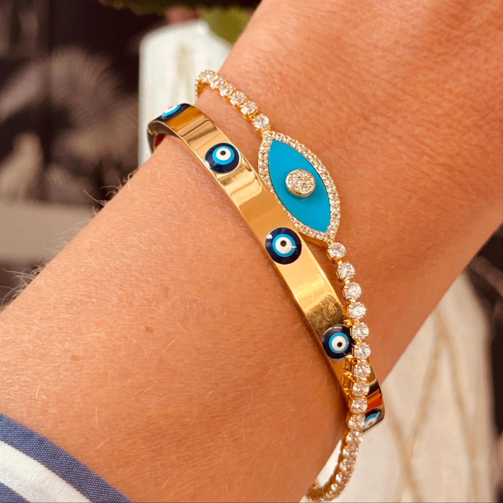 Gold bracelet with blue and black eye design on a wrist, blurred floral background 