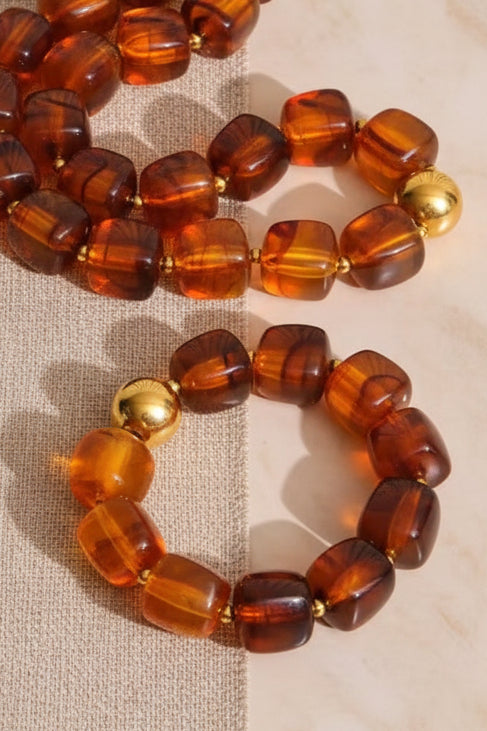 A set of amber beaded jewelry, including a necklace and bracelet, featuring translucent brown and orange beads with polished gold accents, displayed on a light beige stone surface.