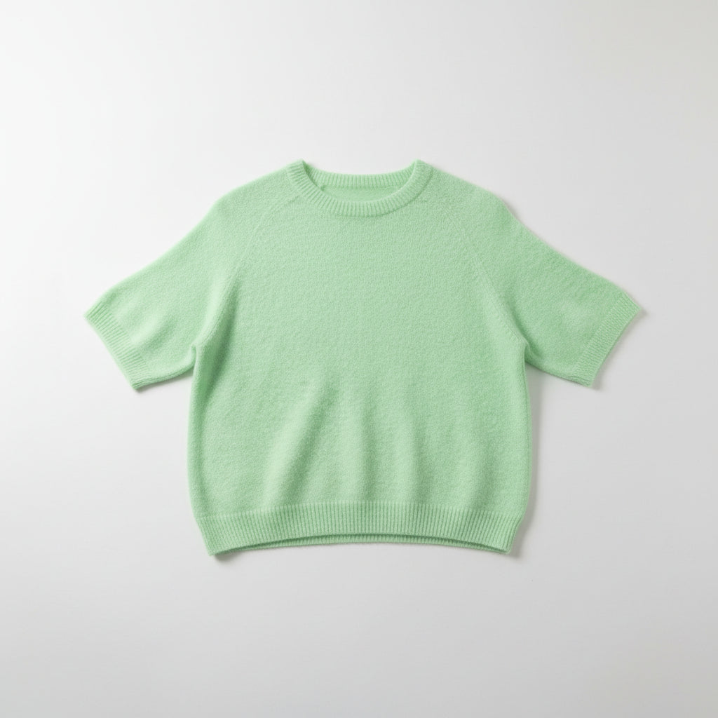 A light mint green short-sleeve knit sweater hanging on a white hanger from a gold clothing rack, set against a gray wood-paneled background.