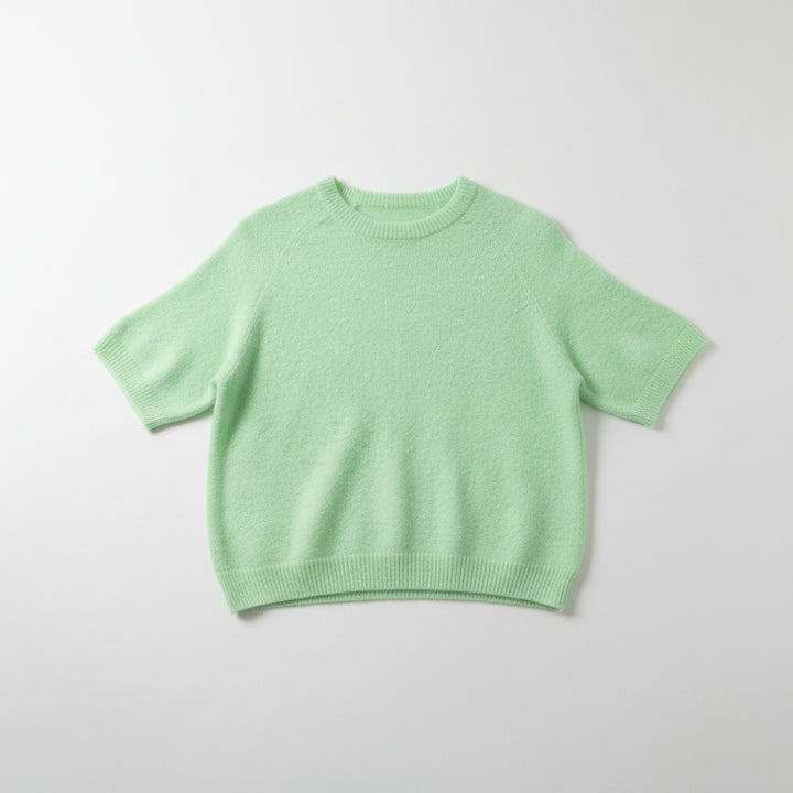 A light mint green short-sleeve knit sweater hanging on a white hanger from a gold clothing rack, set against a gray wood-paneled background.