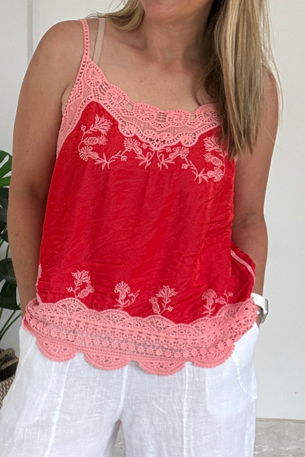 Woman wearing a red top with white lace details and white pants in a room.