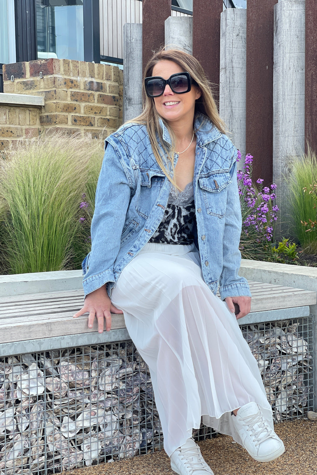 Woman sitting on a bench wearing a denim jacket, white pants, and sunglasses.