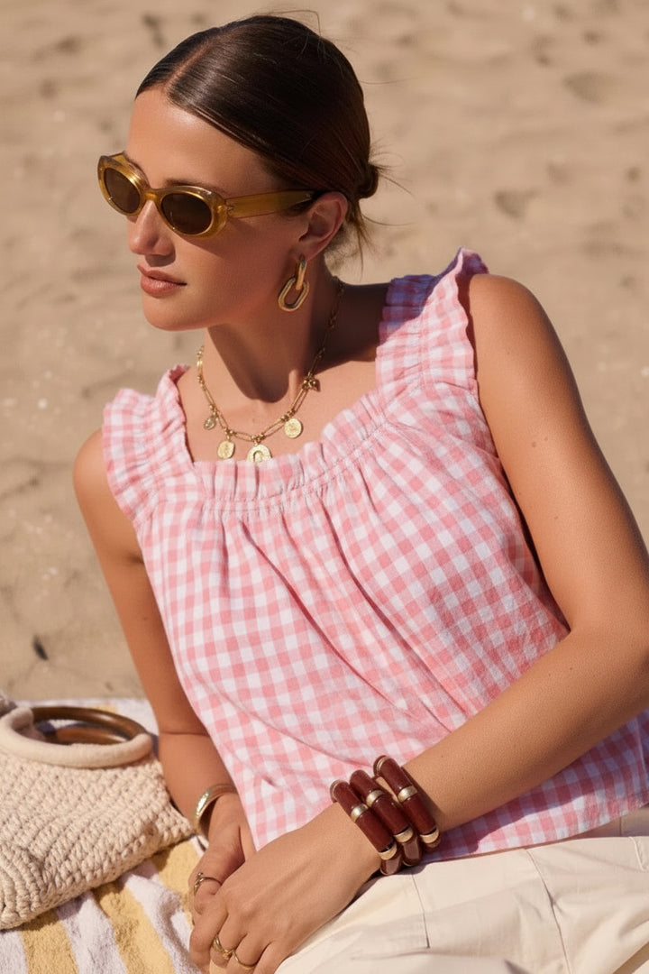 A woman sitting on a beach towel on sand, wearing a red and white gingham sleeveless top, cream pants, gold-framed sunglasses, and layered gold necklaces.