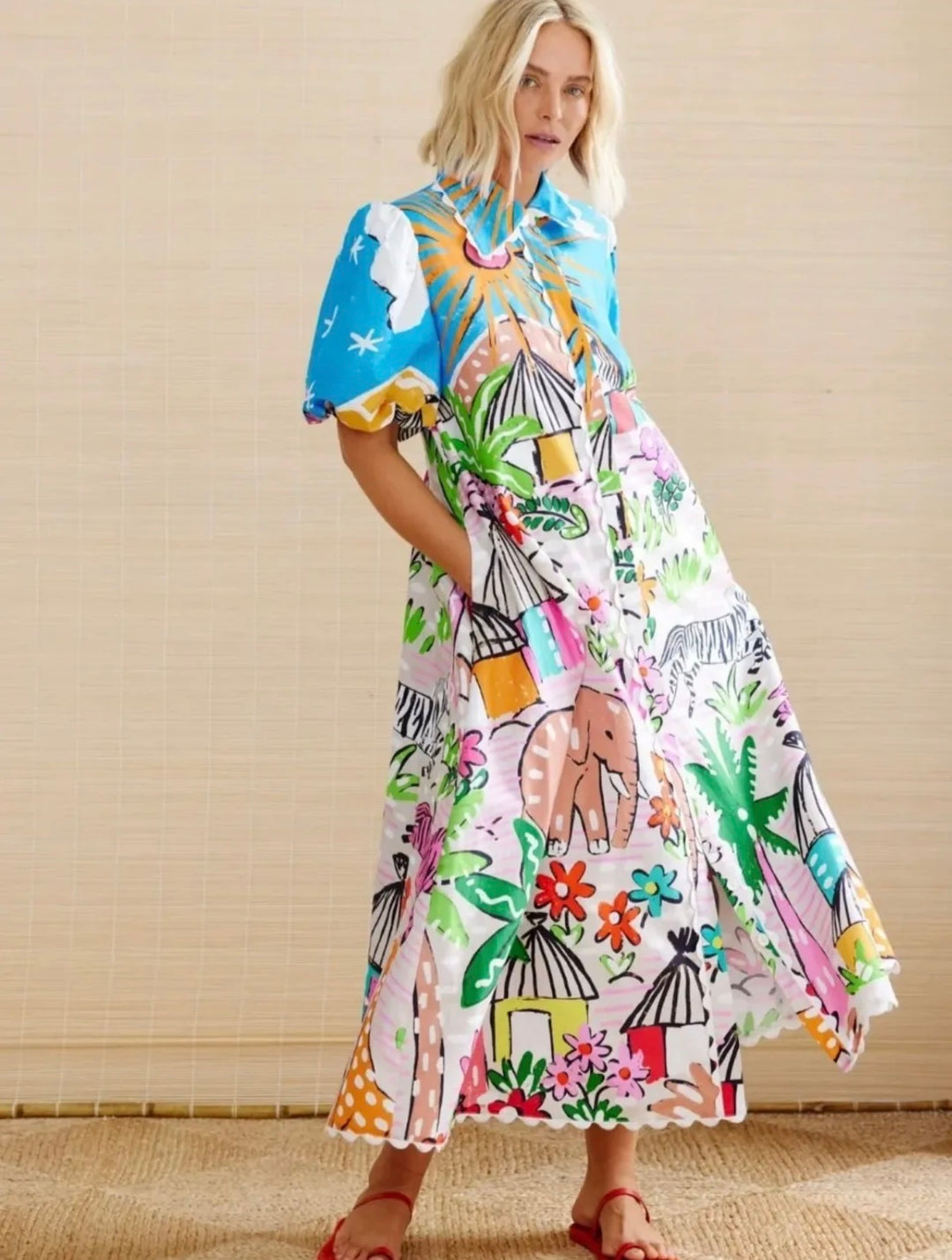 A woman modeling a colorful, short-sleeved maxi dress with a whimsical safari-themed print featuring elephants, palm trees, and tents, set against a neutral beige background.