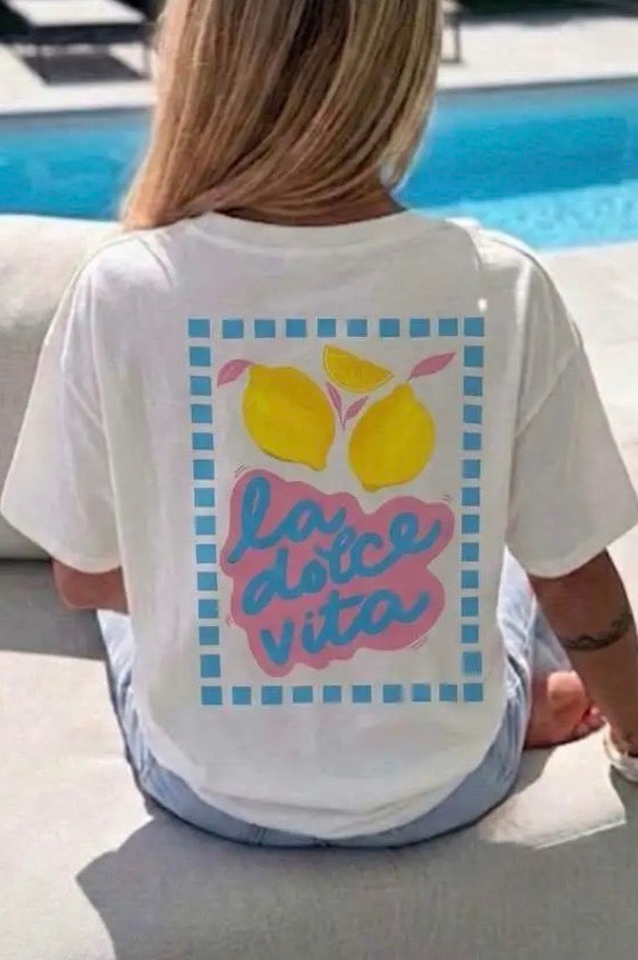 A woman sitting outdoors by a swimming pool, viewed from behind, wearing a white graphic T-shirt with yellow lemons and the Italian phrase 'la dolce vita' in pink and blue on the back.