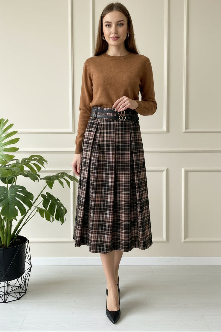 Woman wearing a brown sweater and plaid skirt standing indoors with a plant in the background