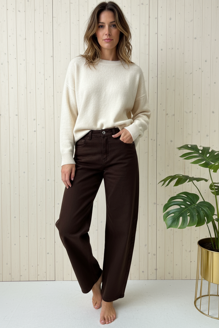 Woman wearing a cream sweater and brown pants standing in a minimalistic room with a plant.