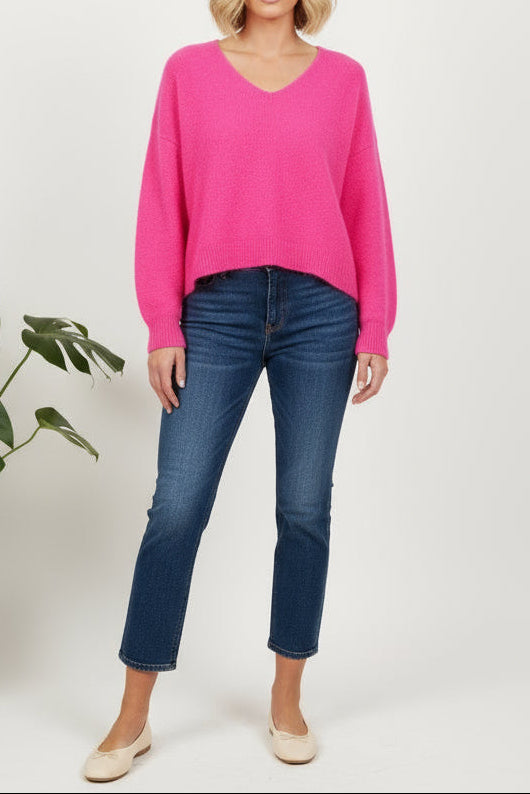 Woman wearing a bright pink sweater and blue jeans standing against a white background with a plant on the left.