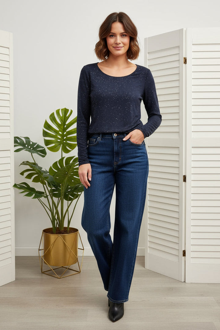 Woman wearing a navy top and blue jeans standing in a room with white walls and a plant.