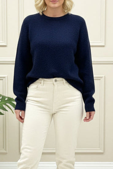Woman wearing a navy sweater and white pants standing in a room with a plant and paneled walls.