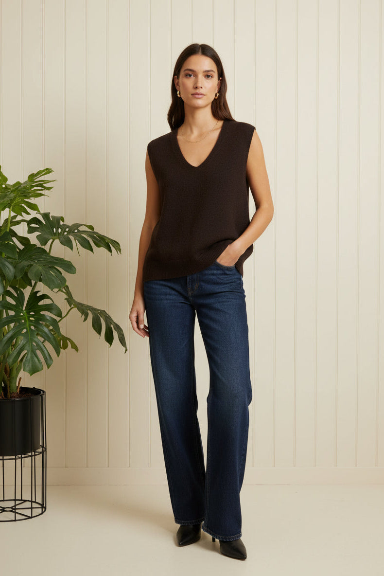 Woman wearing a black sleeveless top and blue jeans standing next to a plant indoors.