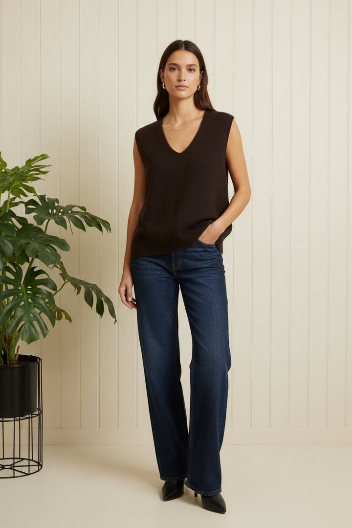 Woman wearing a black sleeveless top and blue jeans standing next to a plant indoors.