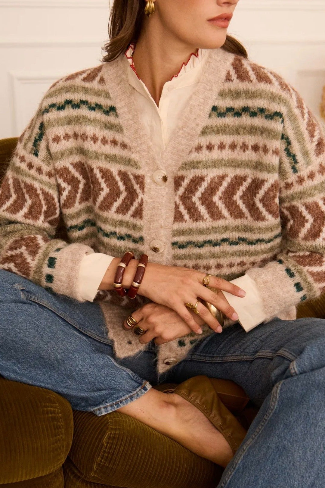 A woman seated on an olive-green couch wearing a fuzzy knit cardigan with a brown, beige, and teal geometric pattern over a white collared shirt and blue jeans.